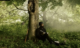 An African American man in an army uniform sits with his back against a tree, writing with a quill pen